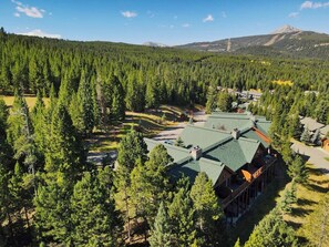 A lodge with a green roof, surrounded by dense forest and mountains in the background. - Perfect Location! High in the Pines, Hot Tub! (Big Sky)