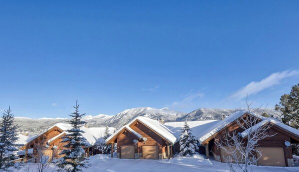 A row of wooden cabins with snow-covered roofs, set against a backdrop of mountains and clear blue sky.