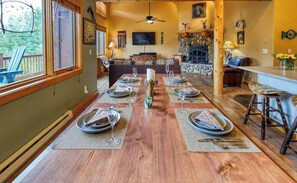 A dining area with a long wooden table set for six, surrounded by chairs, with a stone fireplace and a television mounted on the wall.