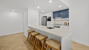 A modern kitchen with a white countertop, stainless steel appliances, and wooden stools. - Luxurious 2BD Ocean View w/ Pool, Gym, Spa & Sauna (Surfers Paradise)