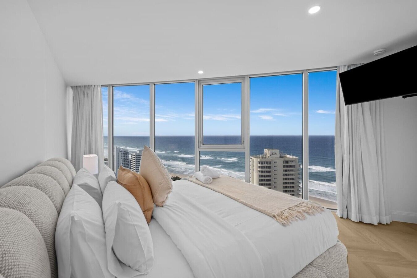 A bedroom with a large window overlooking the ocean, a bed with white bedding, and a view of the sea and buildings.