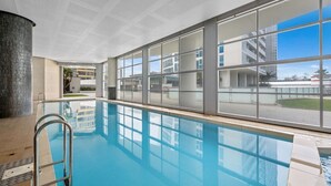 An indoor swimming pool with clear glass walls, a metal ladder, and a view of a modern building complex. - Luxurious 2BD Ocean View w/ Pool, Gym, Spa & Sauna (Surfers Paradise)