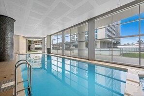 Ein Hallenbad mit großen Glaswänden, einer Metallleiter und Blick auf einen modernen Gebäudekomplex.