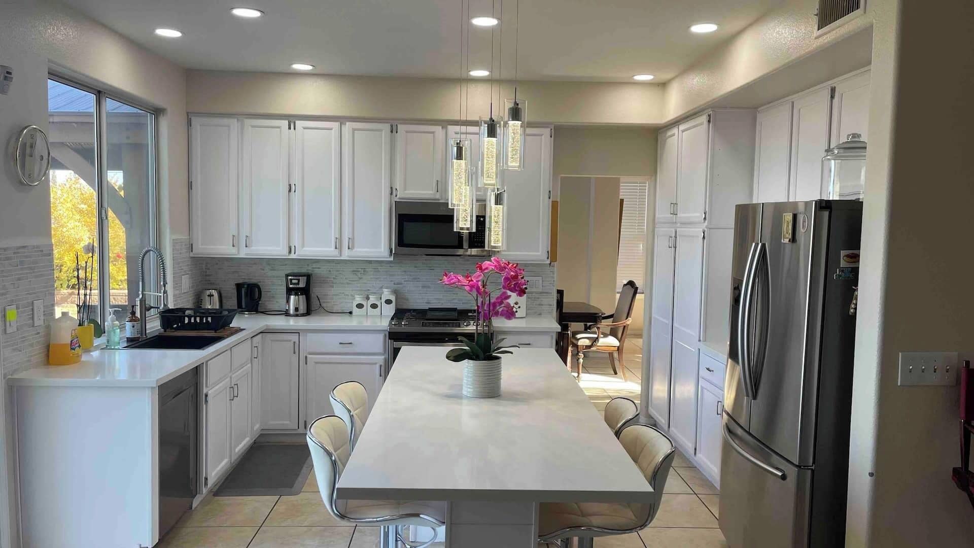 A modern kitchen with white cabinetry, a central island with chairs, a stainless steel refrigerator, and a window providing natural light.