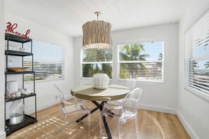 A modern room with a round table, clear chairs, a wooden ceiling, and large windows with blinds. - Sandy Smiles - Gulf View 1-Min-Beach - Heated Pool (Fort Myers Beach)