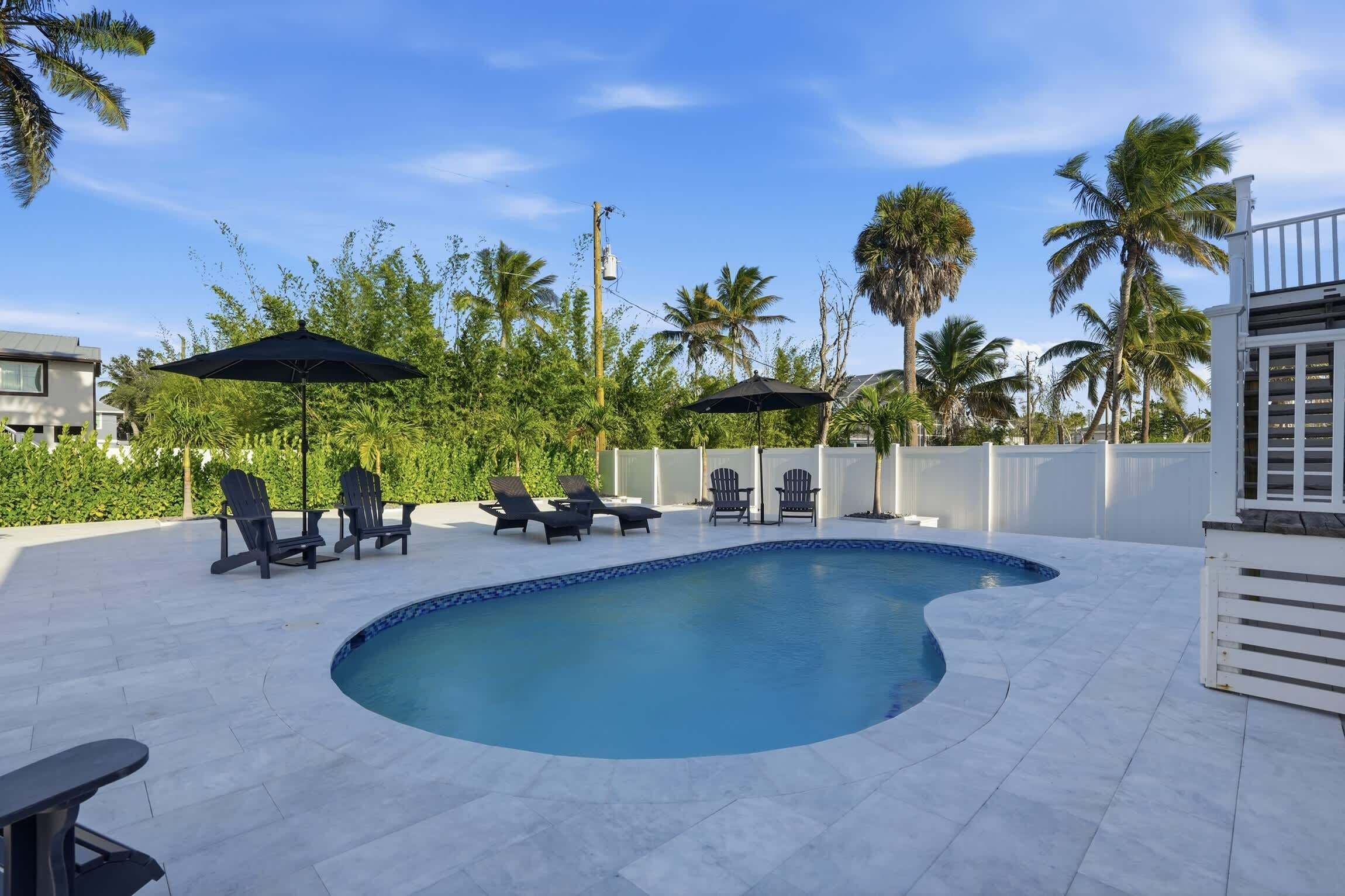 A swimming pool with lounge chairs and umbrellas, surrounded by palm trees and a clear sky.