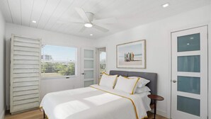 Una camera da letto con un letto, una ventola a soffitto, una finestra con tende, una porta con pannelli trasparenti satinati e un quadro appeso al muro.