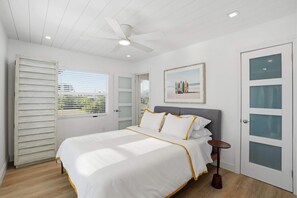 A bedroom with a bed, a ceiling fan, a window with blinds, a door with frosted glass panels, and a framed picture on the wall. - Sandy Smiles - Gulf View 1-Min-Beach - Heated Pool (Fort Myers Beach)