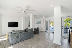 A modern living room with a grey sofa, a flat-screen TV, ceiling fans, and a sliding glass door leading to an outdoor area. - Sandy Smiles - Gulf View 1-Min-Beach - Heated Pool (Fort Myers Beach)
