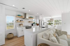 A modern kitchen with white cabinetry, a central island, and a dining area with a white sofa. - Sandy Smiles - Gulf View 1-Min-Beach - Heated Pool (Fort Myers Beach)
