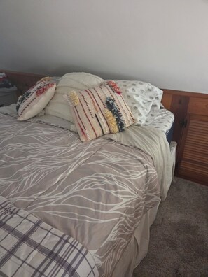 A bed with a patterned bedspread and multiple pillows, a wooden headboard, and a carpeted floor.