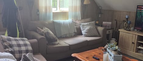 Een gezellige loft woonkamer met een hoge plafond, een houten salontafel, een bank met kussens, een kamerkleed met patroon, en een raam met gordijnen.