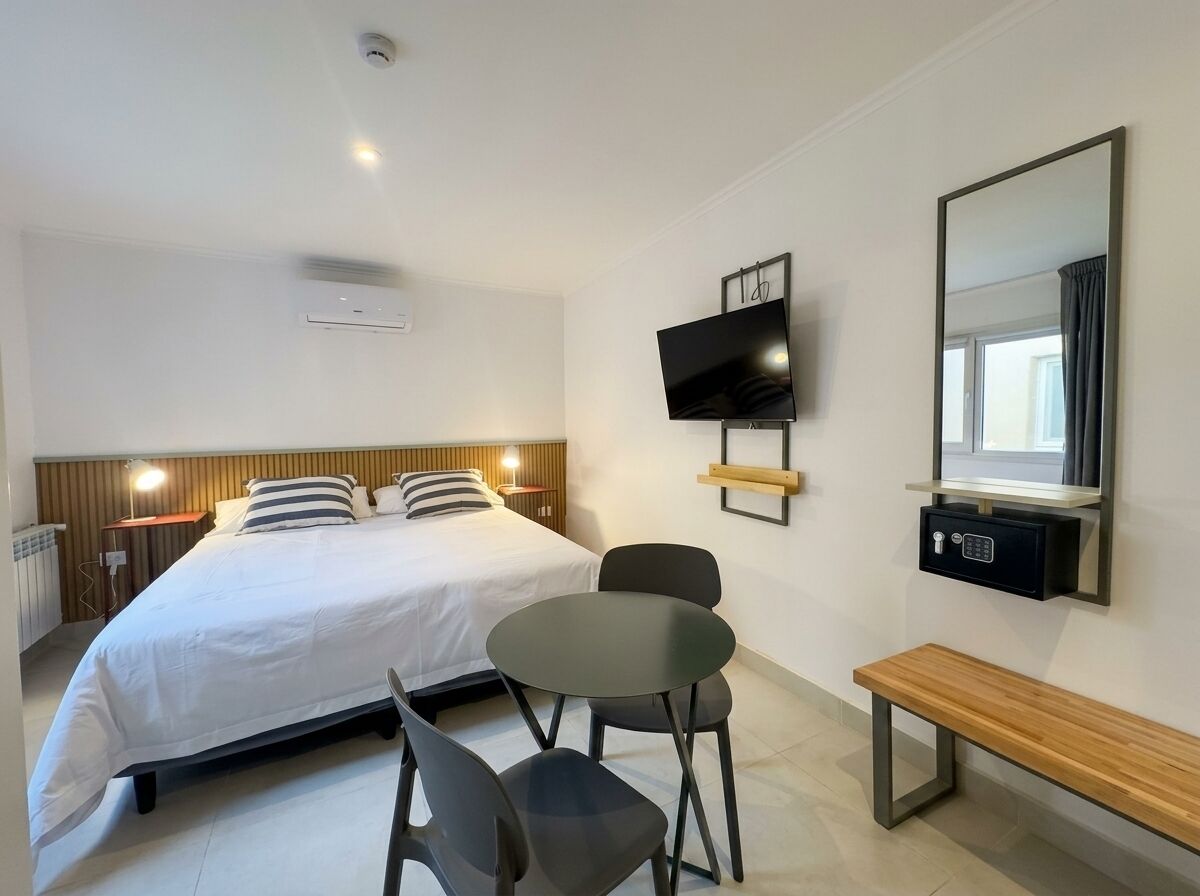 A hotel room with a bed, a TV mounted on the wall, a bedside table, a chair, and a bench.