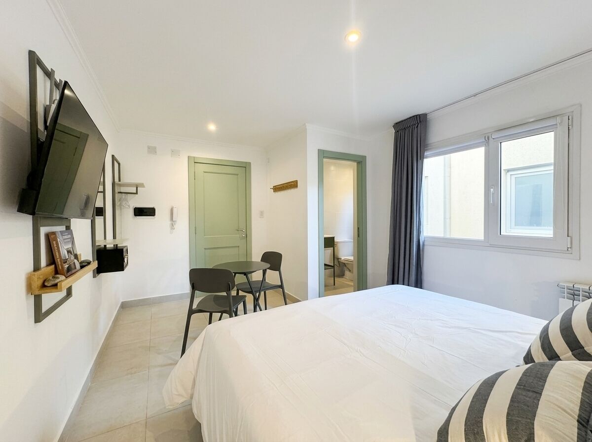 A hotel room with a bed, a TV mounted on the wall, a desk with a chair, and a window with curtains.