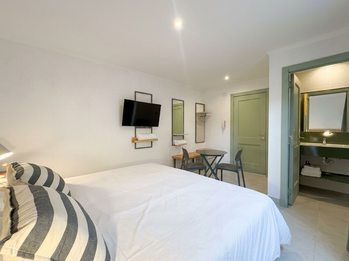 A hotel room with a bed, a TV mounted on the wall, a desk with chairs, and a bathroom with a mirror and sink.