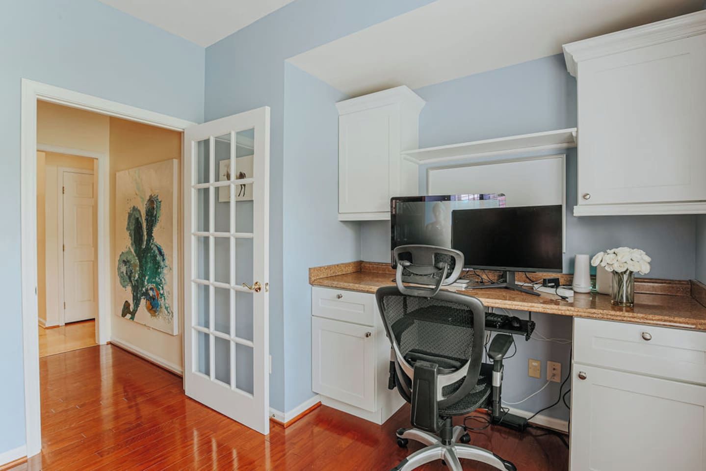Una oficina en casa con escritorio, computadora y silla. Se ve una puerta que da a otra habitación con un cuadro.