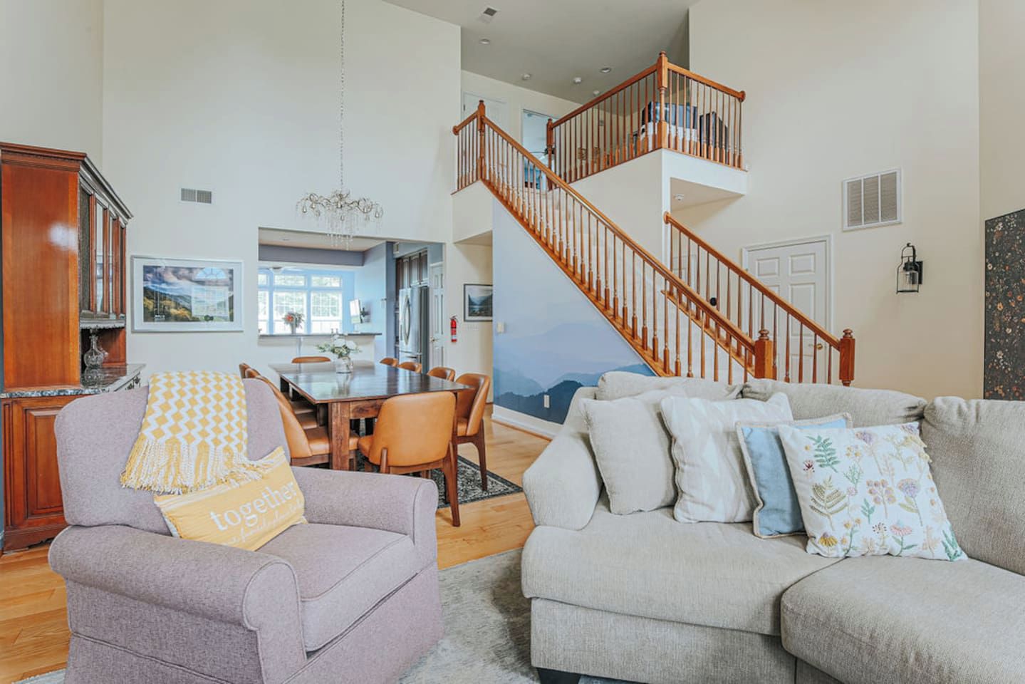 A spacious living room with a high ceiling, a dining area with a wooden table, and a staircase leading to an upper level.