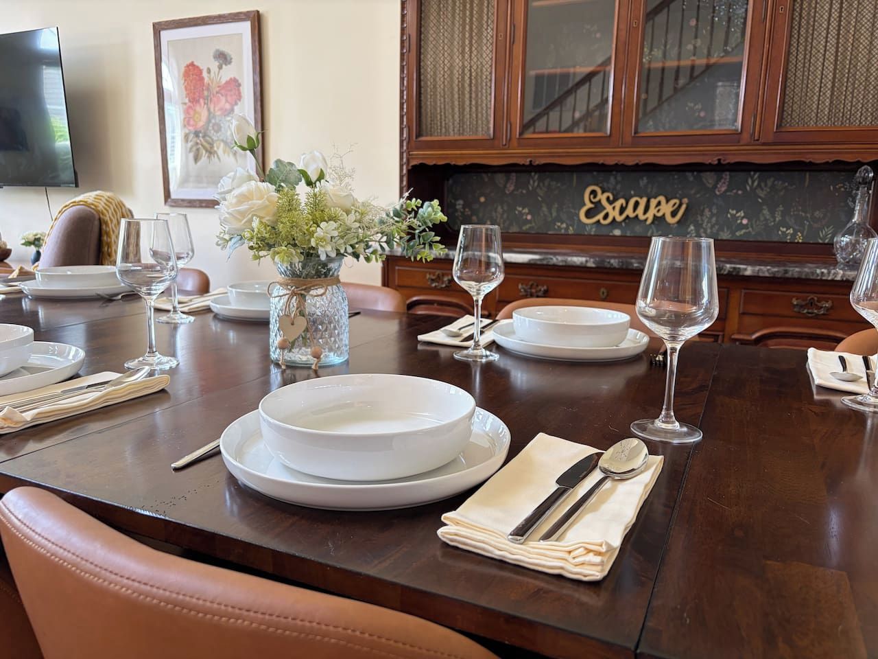 Une table à manger dressée avec des assiettes, des verres et des couverts, un vase rempli de fleurs et un buffet sur lequel est posée une pancarte.