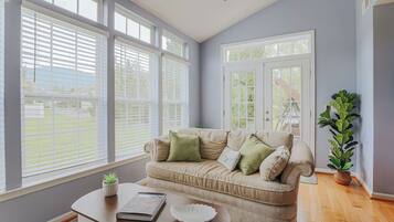 A living room with a beige sofa, a coffee table with a book and a decorative item, large windows with blinds, and a potted plant.