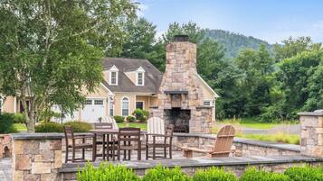 A stone fireplace with outdoor seating, a house with a garage, and a lush green landscape.