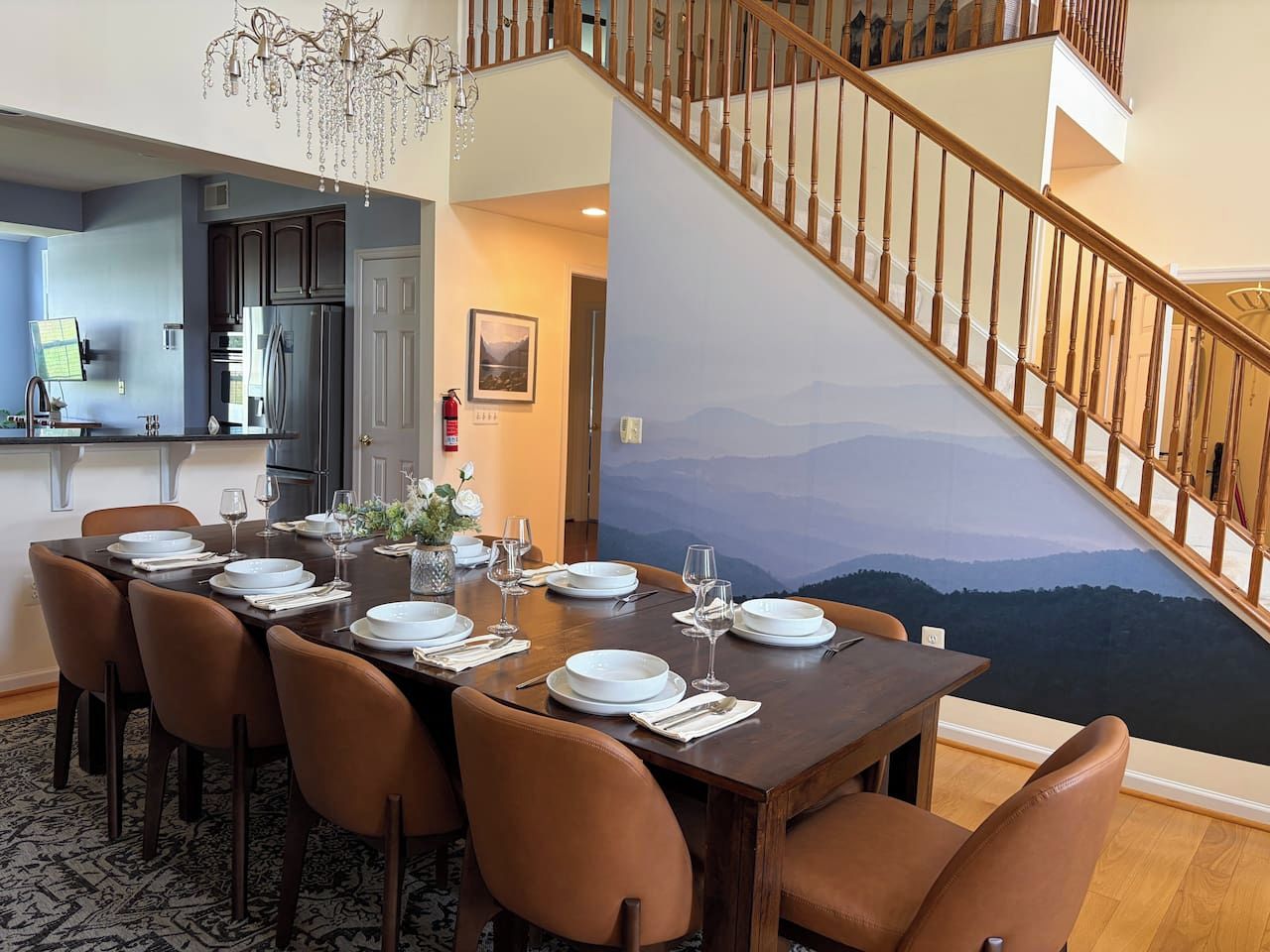 Un coin repas avec une table dressée pour six personnes, une cuisine en arrière-plan et un escalier menant à l’étage supérieur.