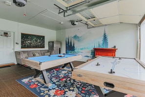 Una habitación con mesa de billar, mesa de hockey de aire, sofá, un cuadro y un mural.