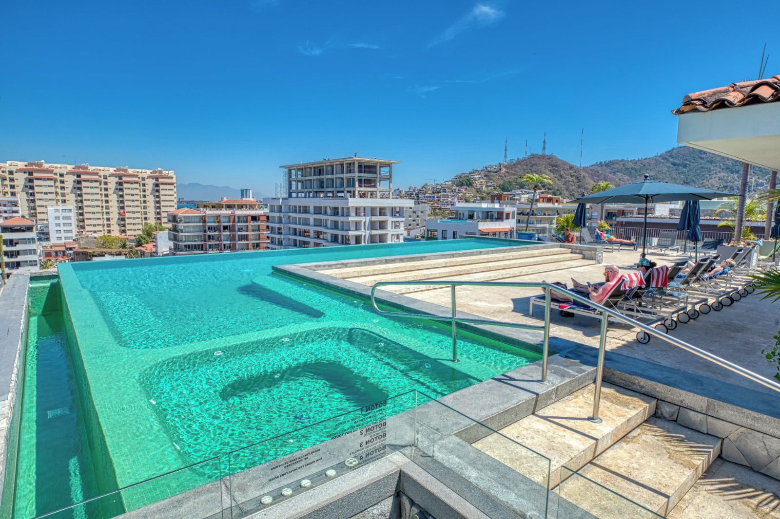 A rooftop swimming pool with clear blue water, surrounded by lounge chairs and umbrellas, with buildings in the background.