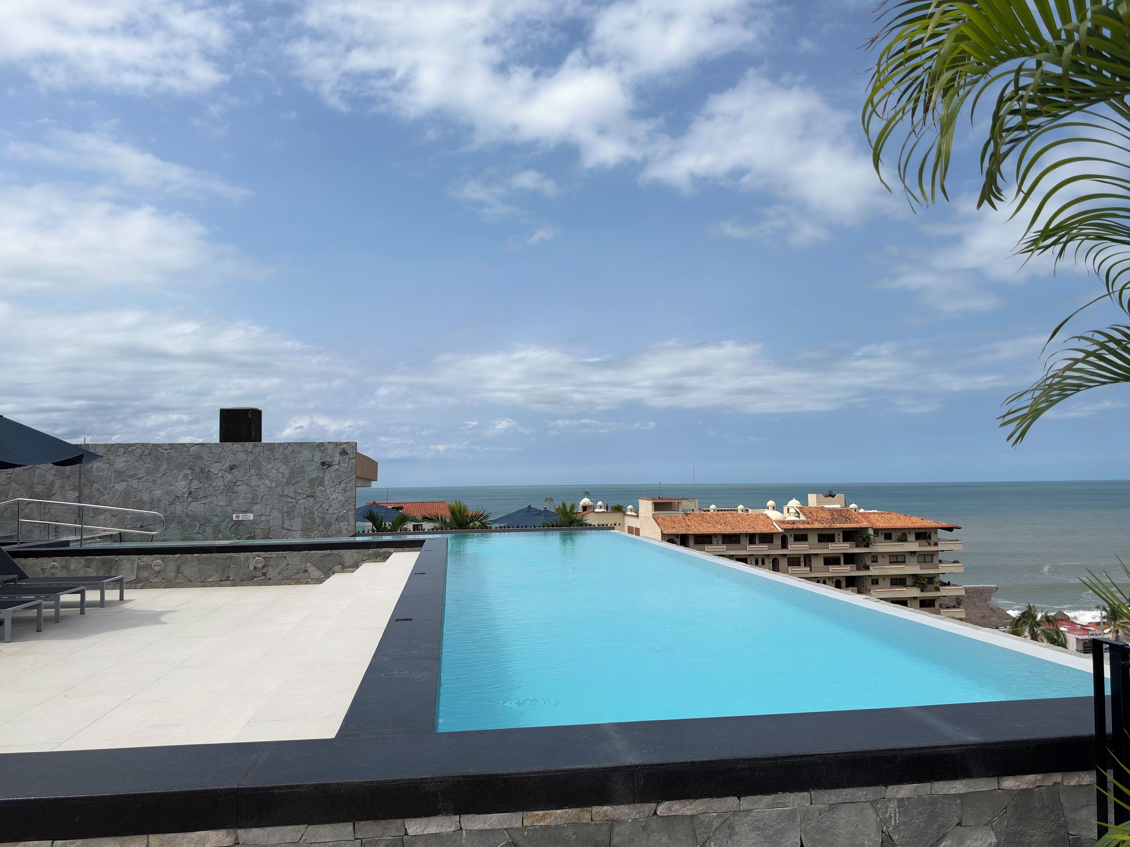 A rooftop infinity pool with a view of the ocean, a stone wall, and a building with a tiled roof.
