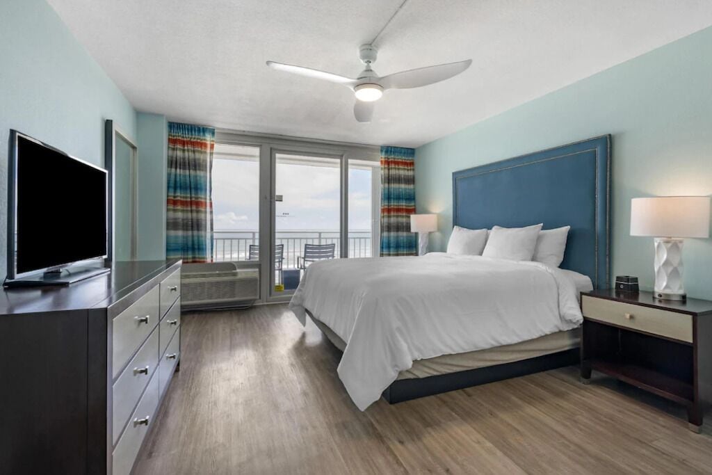 Quarto de hotel com cama, televisão, ventoinha no teto e vista pela janela.