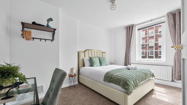 Ein Schlafzimmer mit einem Bett, einem Sessel, einem Schreibtisch, einer Lampe, einer Pflanze, einem Fenster mit Vorhängen und einer Heizung.