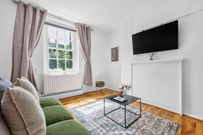 A modern living room with a green sofa, a coffee table, a flat-screen TV mounted on the wall, and a window with curtains. - Cozy Apartment Near Hyde Park (London)