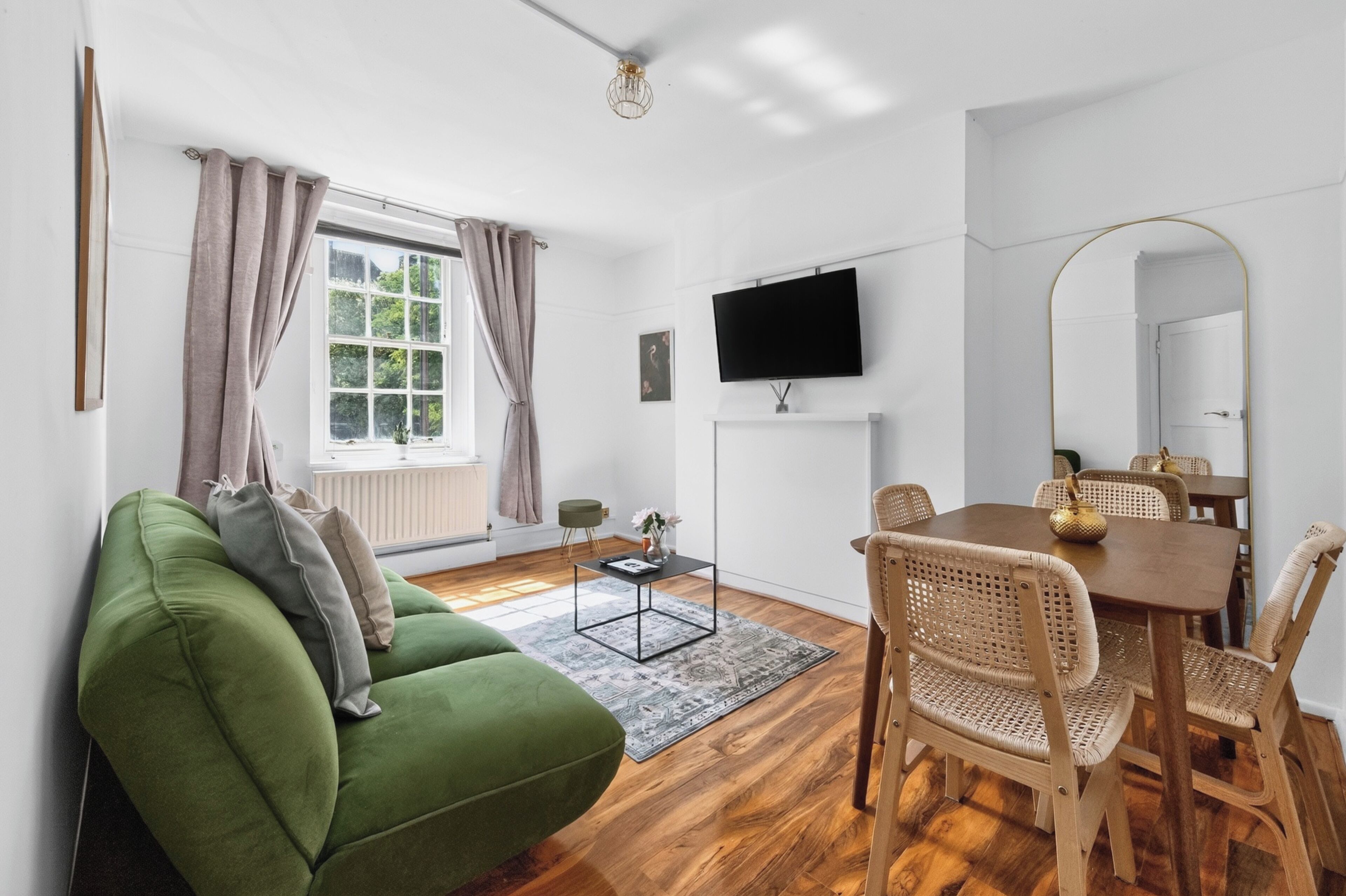 A modern living room with a green sofa, a dining table with wicker chairs, a flat-screen TV mounted on the wall, and a large mirror.