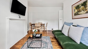 Ein Wohnzimmer mit einem grünen Sofa, einem Couchtisch, einem an der Wand montierten Fernseher, einem Esstisch mit Stühlen und einem Gemälde an der Wand.