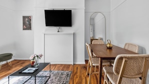 Ein modernes Wohnzimmer mit Flachbildfernseher an der Wand, einem Esstisch mit Korbstühlen, einem Couchtisch mit Blumenvase und einem gerahmten Bild.