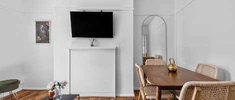 A modern living room with a flat-screen TV mounted on the wall, a dining table with wicker chairs, a coffee table with a vase of flowers, and a framed picture on the wall.