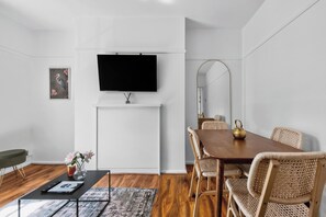 A modern living room with a flat-screen TV mounted on the wall, a dining table with wicker chairs, a coffee table with a vase of flowers, and a framed picture on the wall. - Cozy Apartment Near Hyde Park  (London)