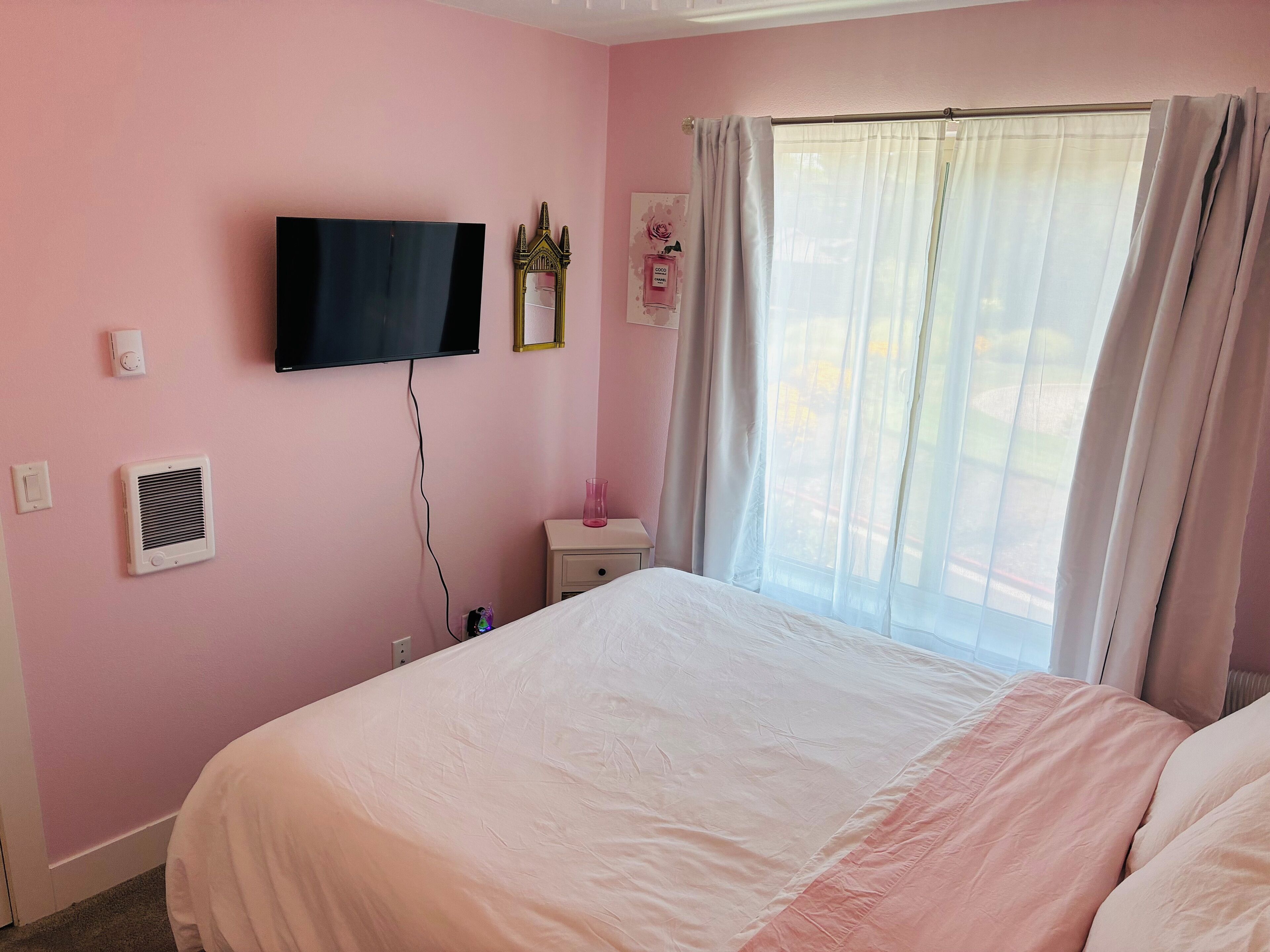 A bedroom with a bed, a television mounted on the wall, a bedside table with a lamp, and a window with curtains.