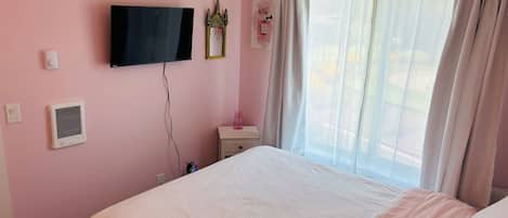 A bedroom with a bed, a television mounted on the wall, a bedside table with a lamp, and a window with curtains.
