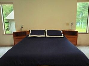 Una cama con una colcha azul oscuro y dos almohadas, flanqueada por mesitas de noche de madera con lámparas, en una habitación con ventanas que dan a vistas de vegetación.