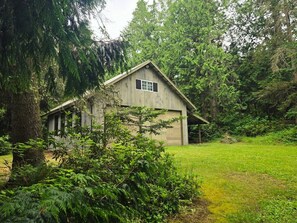 A wooden cabin with a gabled roof, surrounded by dense greenery and trees. - Tranquil Bluff retreat W/ Water Views & Game Room (Marrowstone)
