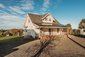 A two-story house with a covered porch, a gravel driveway, and a fenced yard. - Farm Cottage in Sequim w/ Fenced Orchard (Sequim)