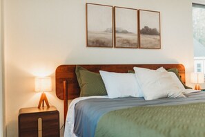 Una habitación de hotel con cabecera de madera, una cama con muchas almohadas, una lámpara de noche y tres cuadros en la pared.