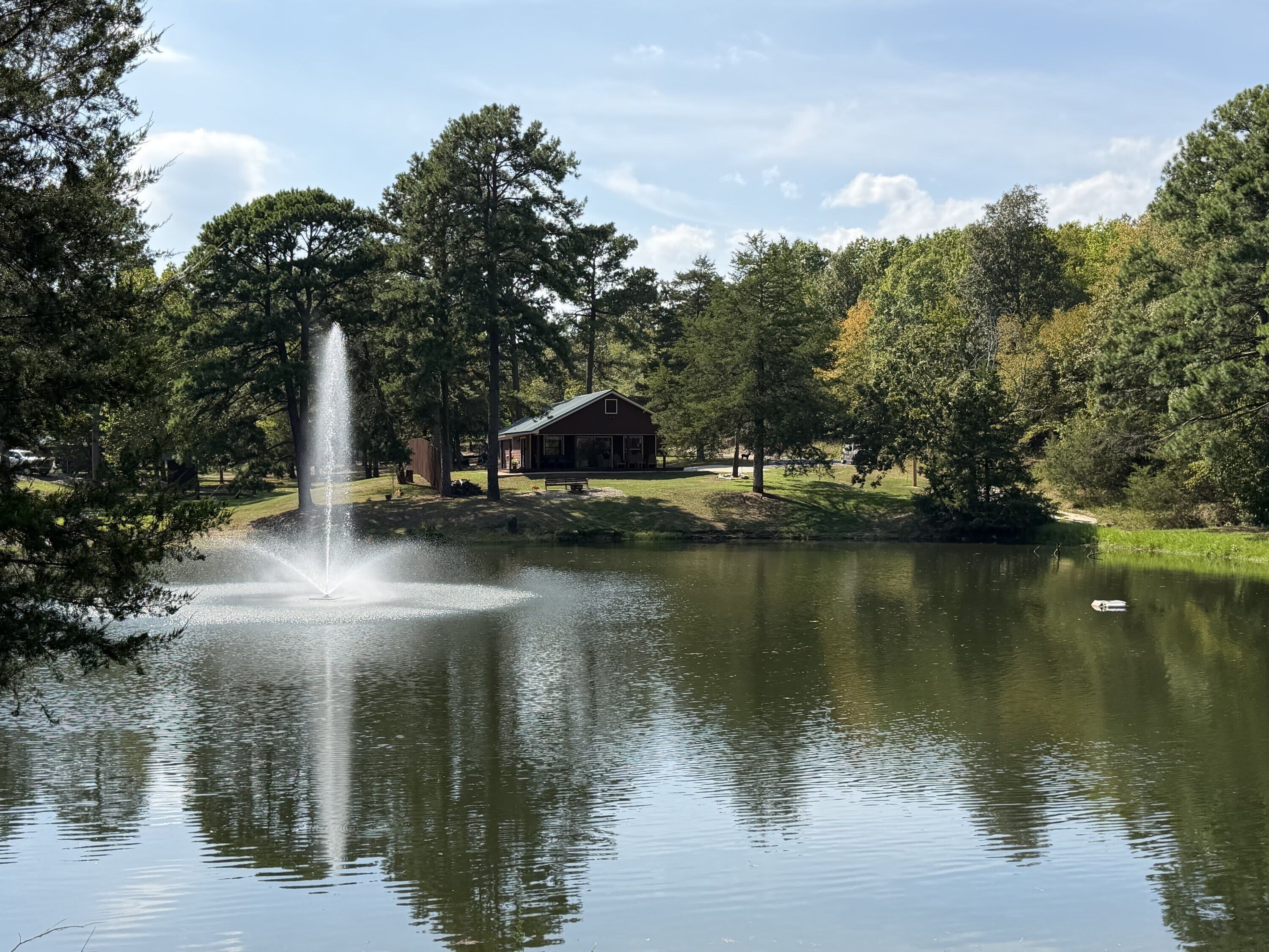 Un lago tranquilo con una fuente, rodeado de árboles y una cabaña.