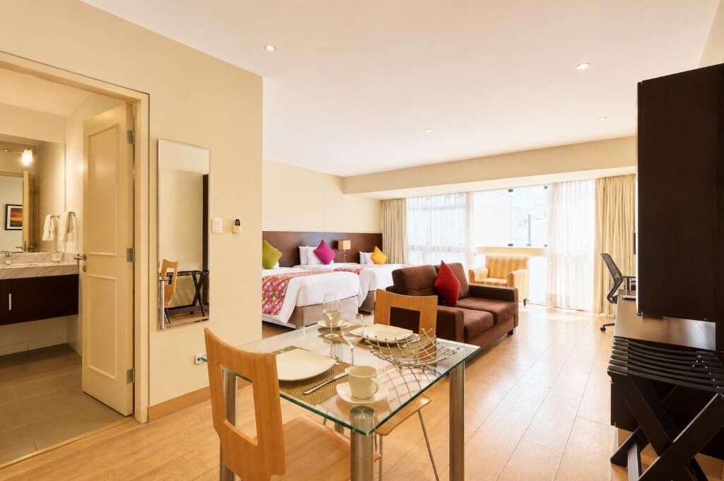 Habitación de hotel con zona de comedor, cama con almohadas coloridas, televisor y ventana grande.