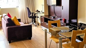 A modern living room with a sofa, a glass dining table, a TV mounted on the wall, and a kitchen area with a stove. - Diez Canseco 2W Apartments Service (Miraflores)