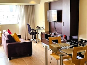 Una sala moderna con un sofá, una mesa de comedor de vidrio, un televisor montado en la pared y una zona de cocina con estufa.