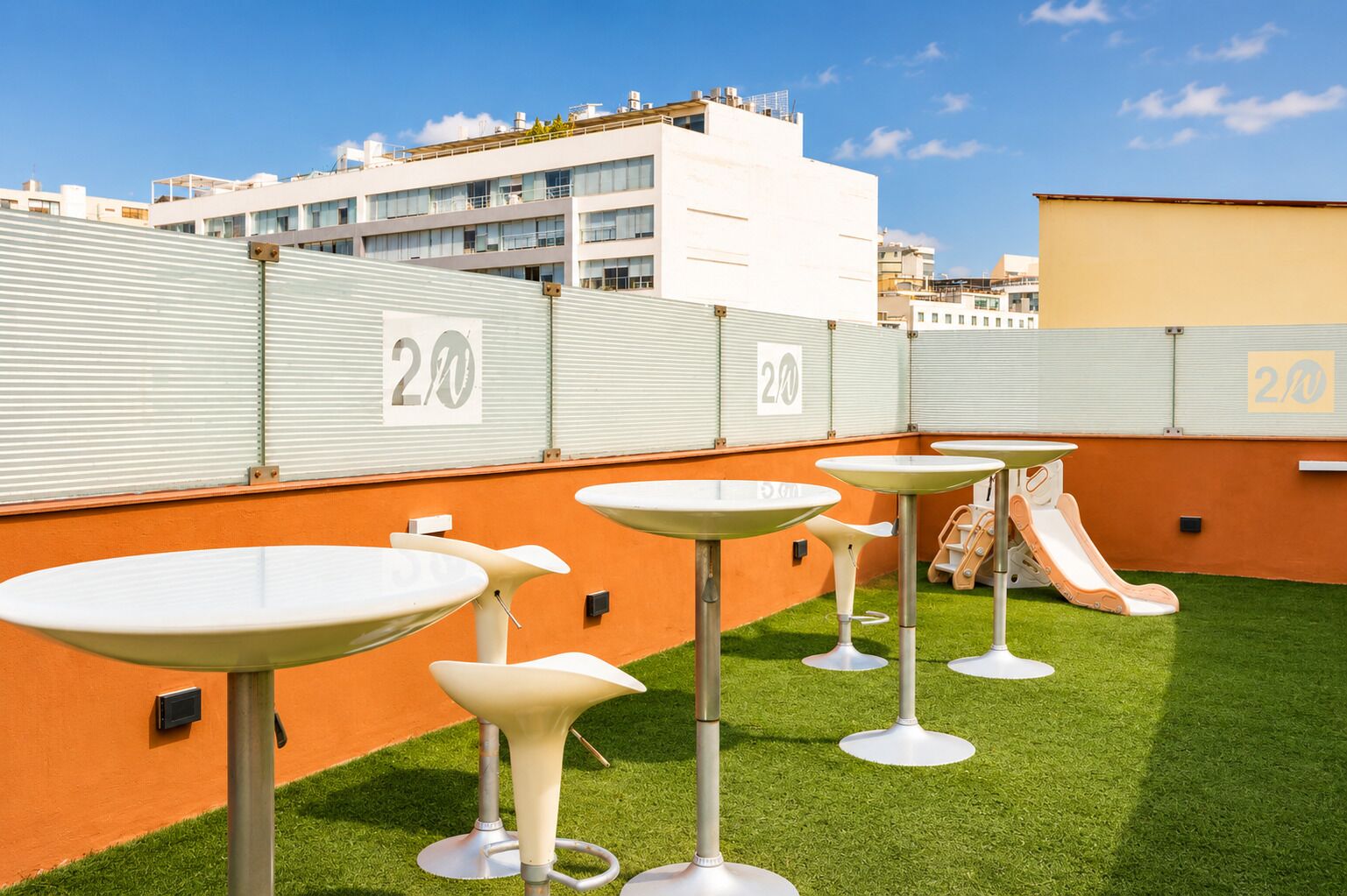 A rooftop terrace with multiple white tables and a slide, surrounded by artificial grass and a metal fence with the number '20' on it.