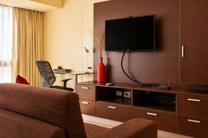 Une chambre d’hôtel équipée d’un téléviseur à écran plat fixé à un meuble en bois, d’un bureau en verre avec une chaise, et d’un vase rouge contenant des plantes séchées.