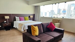 A hotel room with a bed, a sofa, a nightstand, and a window with a city view. - Diez Canseco 2W Apartments Service (Miraflores)