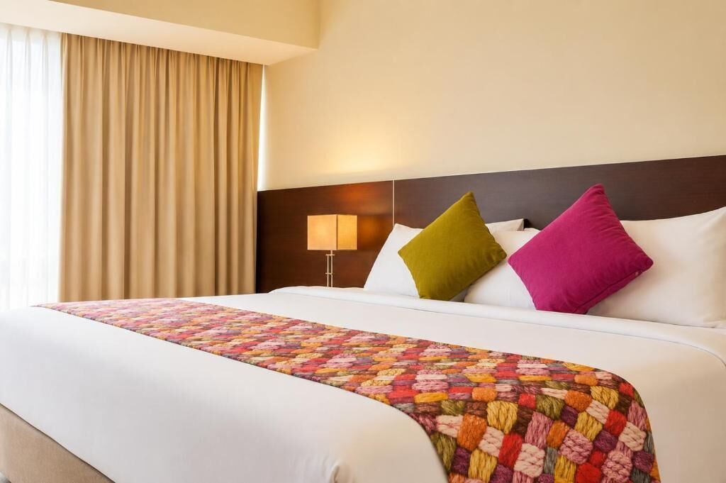 Habitación de hotel con una cama grande, cabecero, lámpara de noche y ventana con cortinas.